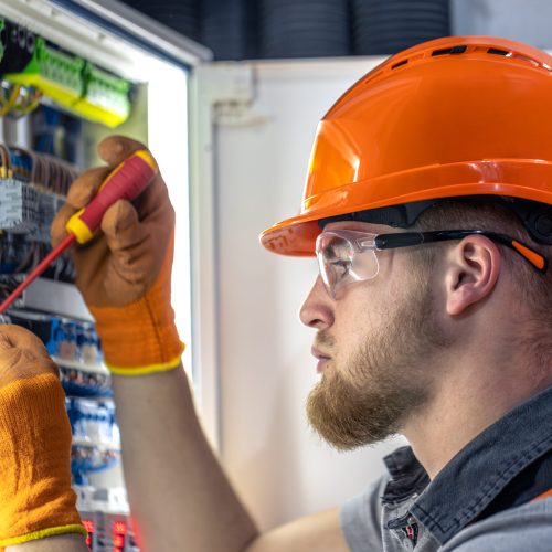 Male electrician working in switchboard. Male electrician in overalls working with electricity. High quality photo. Close-up. Man wearing safety glasses and hard hat.