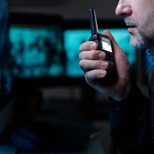 Hand of mature male guard in black uniform speaking in walkie-talkie and looking at computer screen while sitting in front of monitor