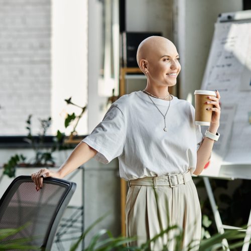 A young bald woman in elegant dress stands by a chair, smiling with a coffee cup in an office.